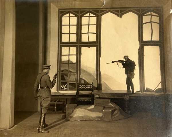 A production photograph. The stage set is a grand largely empty room with a solid wooden chair and table. Two men dressed as soldiers reminiscent of the  First World War stand on opposite ends of the stage points guns at one another