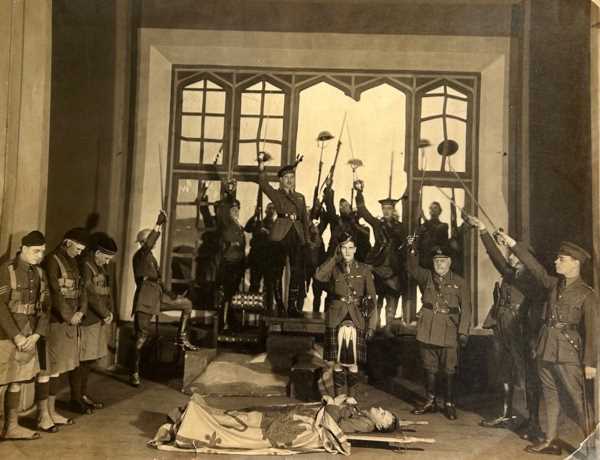 A production photograph. The stage set is a grand largely empty room with a broken window to the read. Many men dressed as soldiers reminiscent of the First World War stand on opposite ends of the stage, including some in kilts, raise swords in salute to a man lying on a stretcher, apparently dead draped in a heraldic flag.