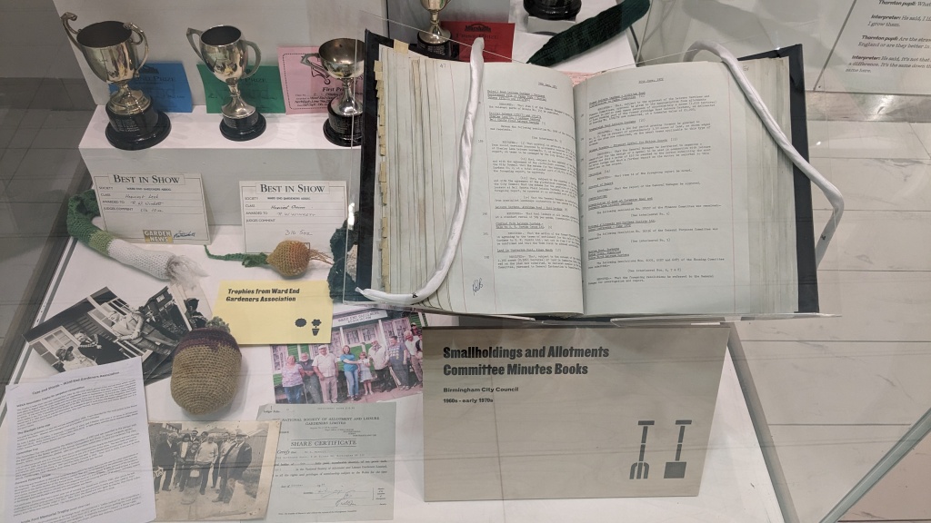 A close up of on efo the exhibtion case. A BCC minute book is held opne, other items in the box include trophies and best in show certificates, photographs of groups of allotmenteers and knitted vegetables