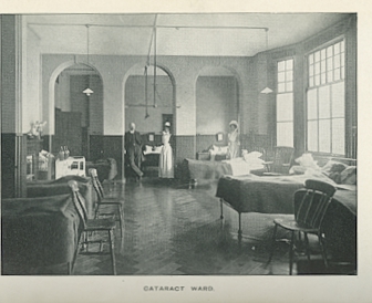 Airy room with large windows and 6 beds arranged symmetrically. a Man in a suit is standing in the hallway and there are two nurses with long white aprons over their full floor length dresses.