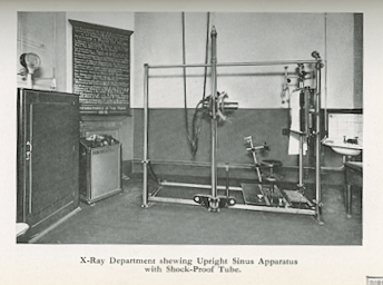 Black and white photograph showing the corner of a room full of equipment, there is a black board and a sink and a large metal stand which appears to have a camera on it