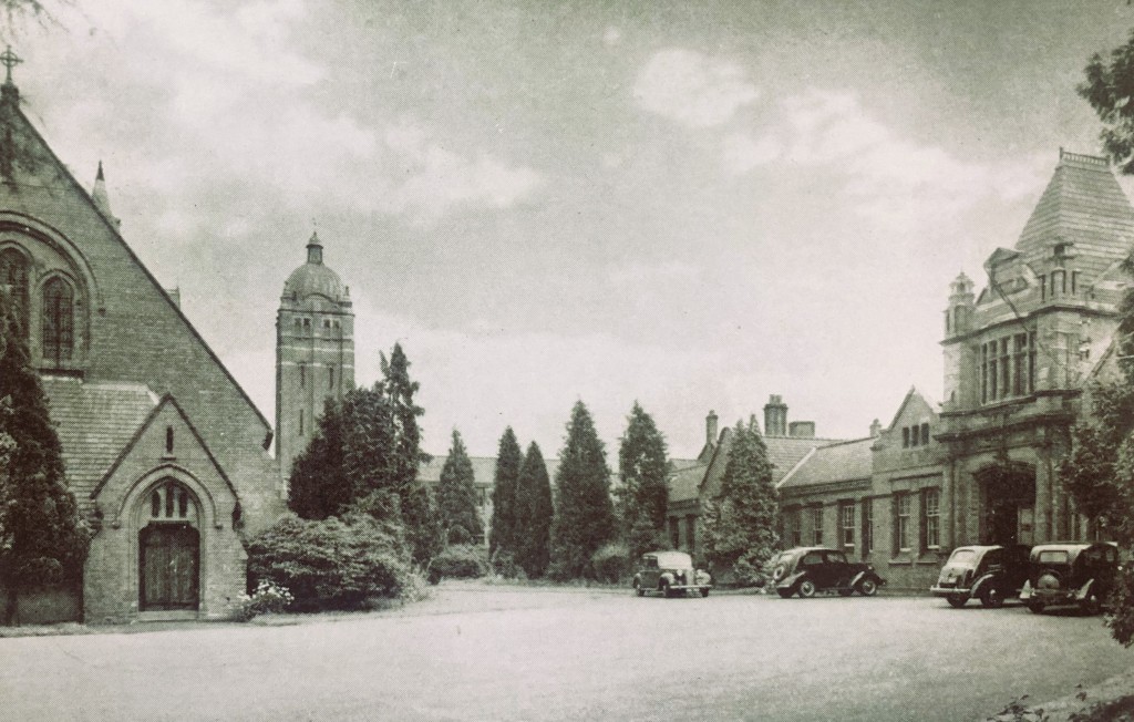 Hollymoor Hospital's early 20th century brick buildings with conifers and 1950s cars parked outside the entrance on the right, with the hospital chapel on the left. Image in black and white. 