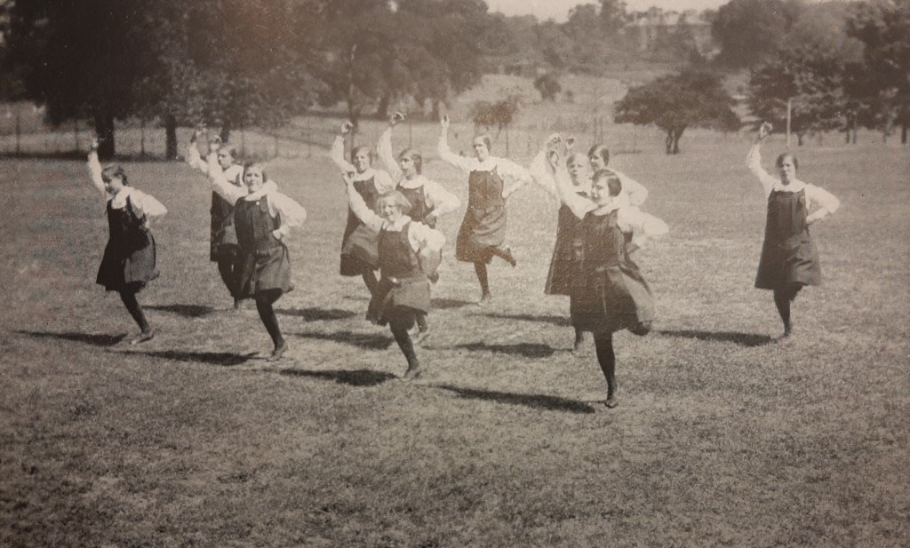 Group of 11 girls from the Royal School for Deaf Children in landscaped grounds dancing with one hand on their hips and one arm in the air. They are dressed in dark coloured knee-length pinafore dresses with white blouses, dark stockings and shoes.