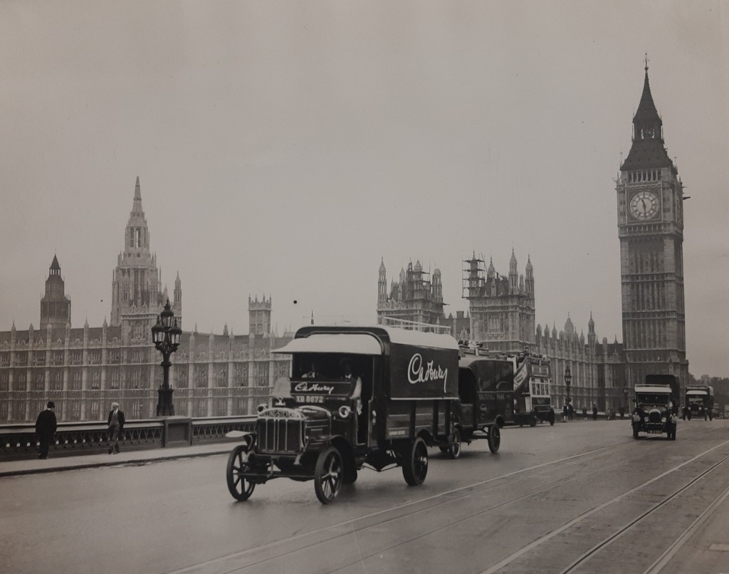 1920s vehicles, including a Cadbury's delivery van, driving along a road bridge with the Palace of Westminster in the background. 