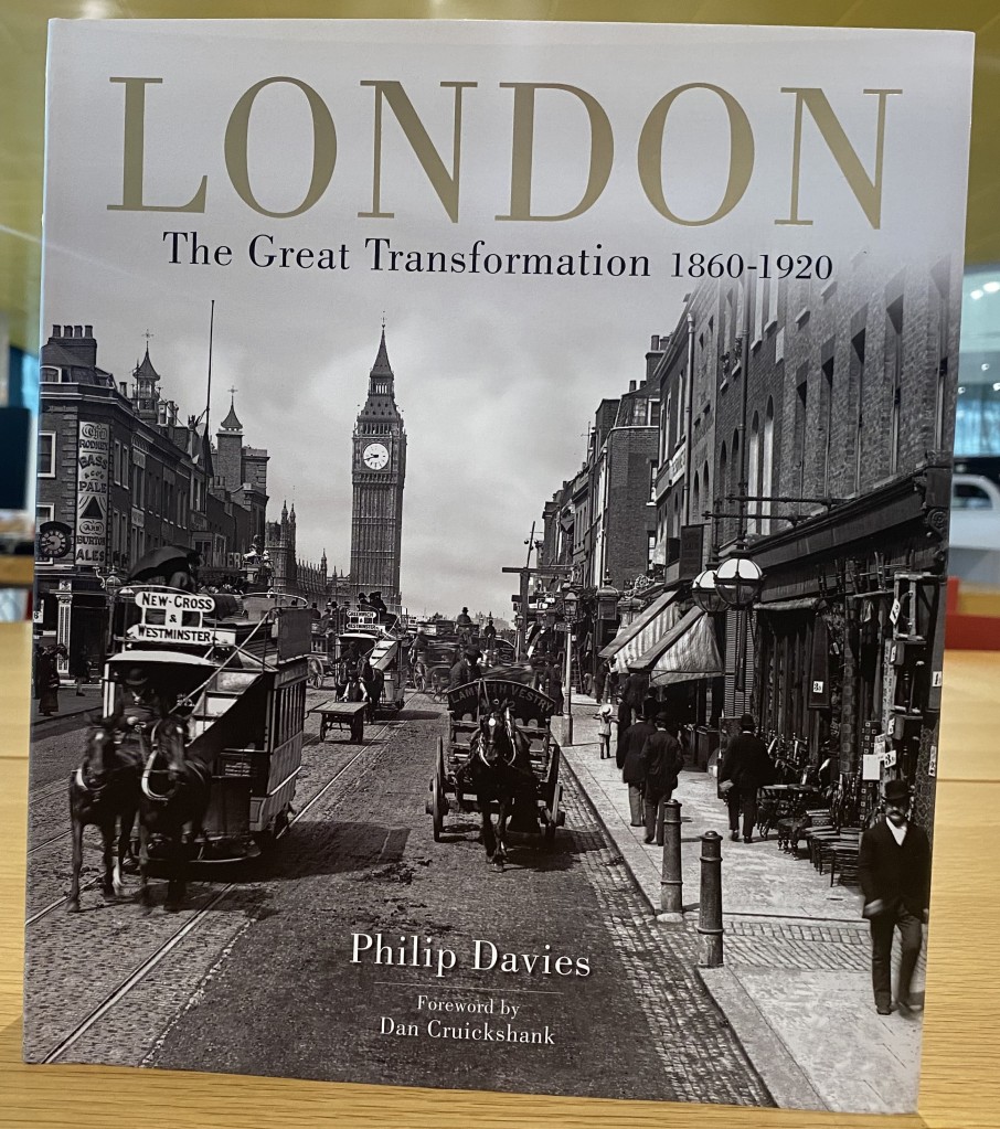 Black & white cover of a book showing a 19th century street scene in London with Big Ben in the background. The book is set upright on a wooden table. 