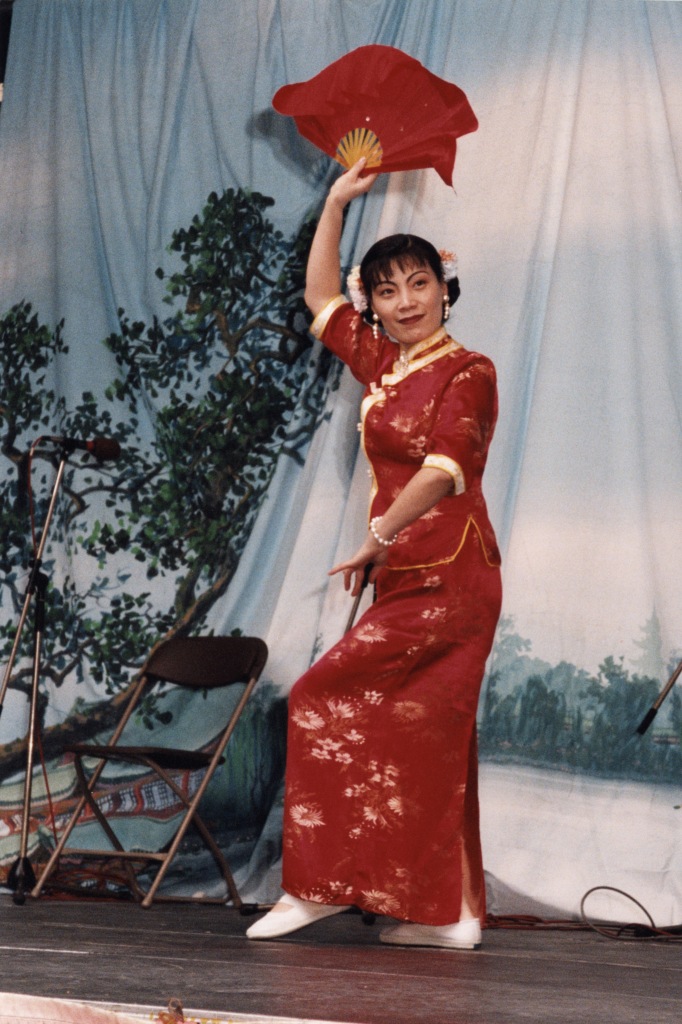 Person dancing in red and gold traditional red dress ( skirt and top) the outfit is bordered in gold with gold flower design on the fabroc. The dancer wears floweres in her hair and is holding fan above her head