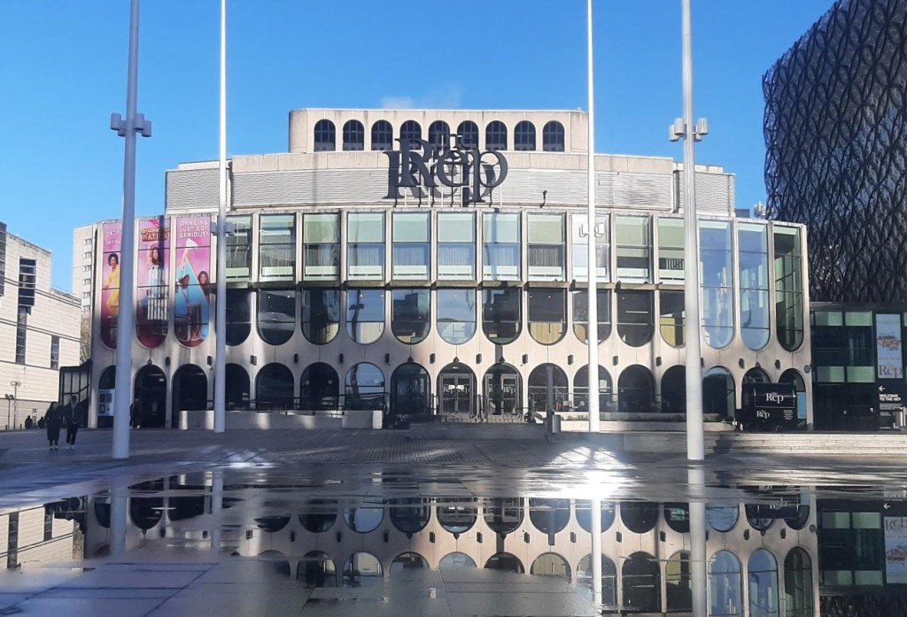1960s light-coloured theatre building with glass facade looking onto a town square which has reflections of the building in puddles. The square as 4 flag poles.