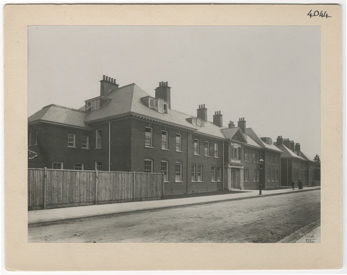 A large brick building with many windows and chimneys, with fences to the left of the image and an empty road in front of it.