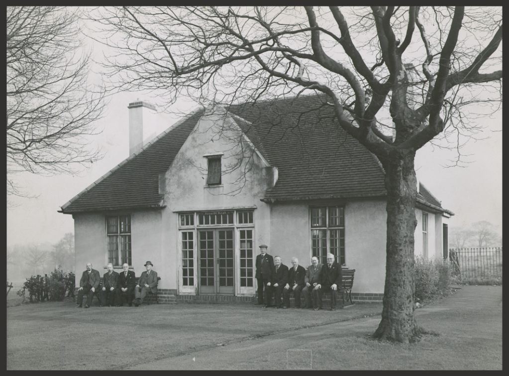 9 men in suits sit on benches in front of a two storey detached cottage or pavilion style building which has white washed walls and large windows.
