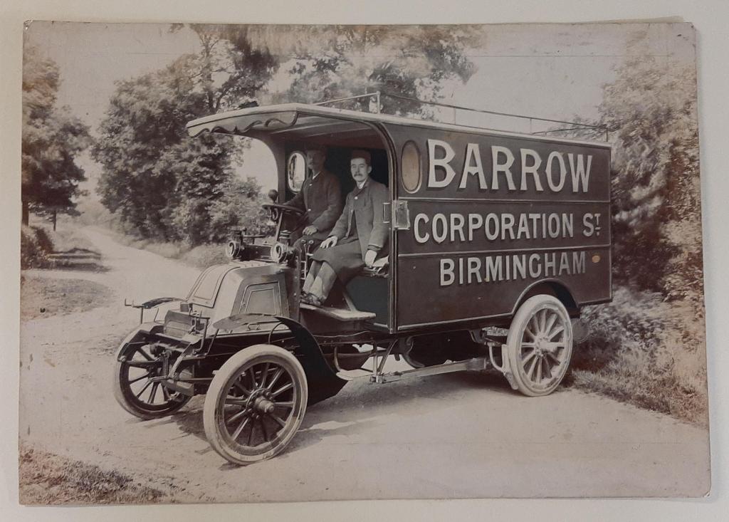 A 1920s delivery van with two men seated in it, parked on a country road lined with trees