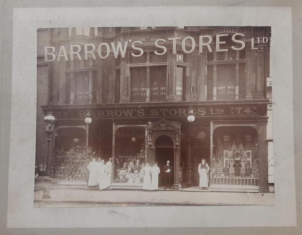 Victorian shop front with staff dressed in white aprons standing outside and shop windows displaying goods