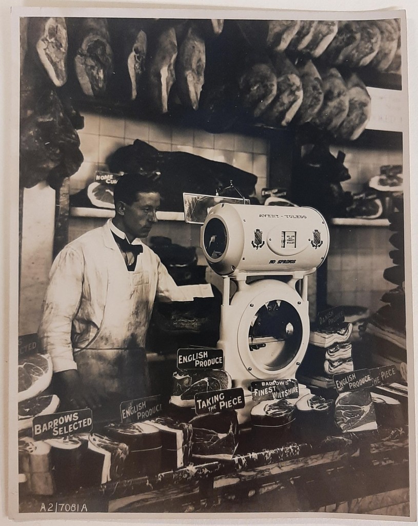 A man dressed in white behind scales at a cooked meat counter in a shop.