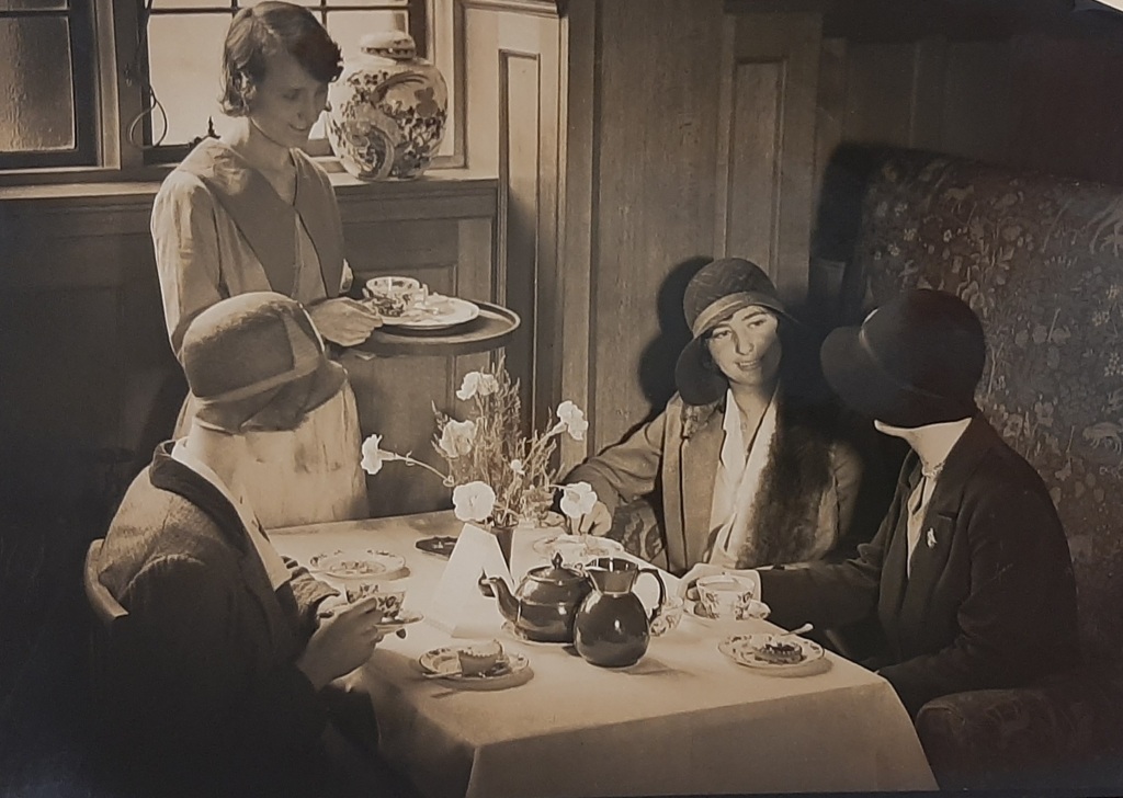 Three smartly dressed women in cloche hats seated at a table which has a tea pot, tea cups, a vase of flowers and cake  on it. A waitress is standing by the table with a tray in her hand, about to serve a cup of tea.