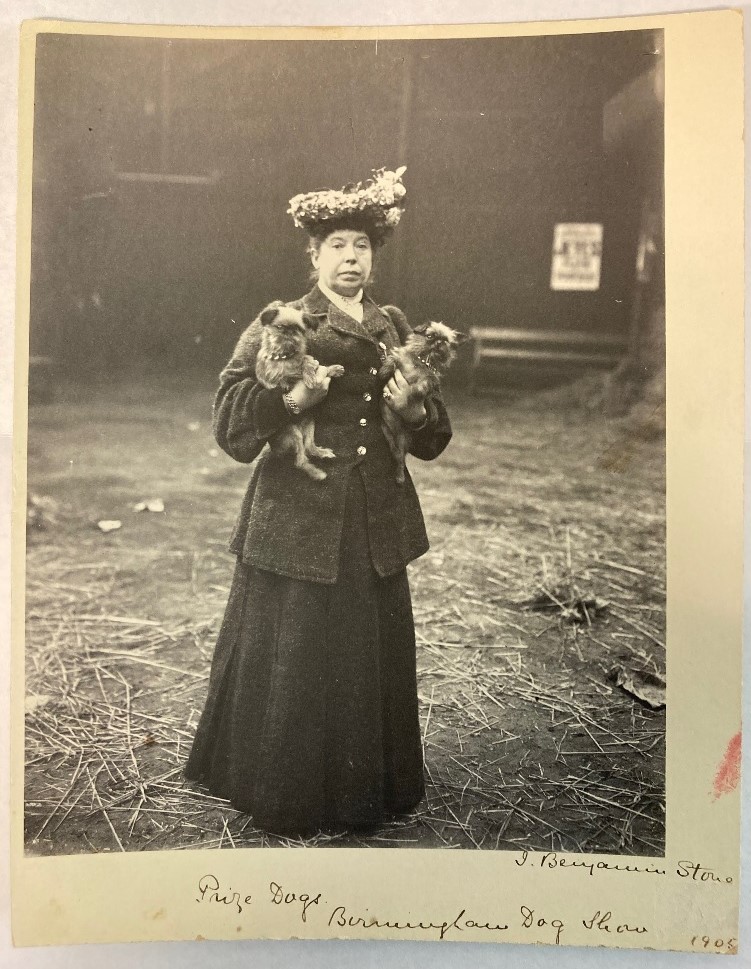 Full length portrait of a woman in Edwardian dress, wearing a full length skirt, high necked blouse and  coat with a large hat adorned with flowers, holding a small dog in each arm. There is straw on the ground. 