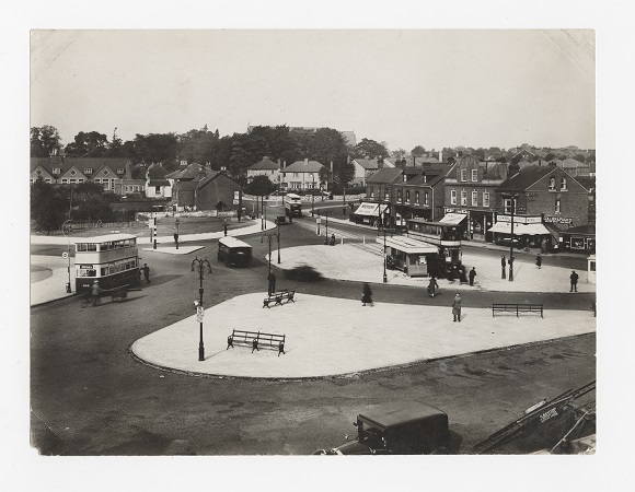 A photograph of Acocks Green Village taken in 1928 showing a village green with benches, and bus stops. Buses and shops are also visible in the image. 